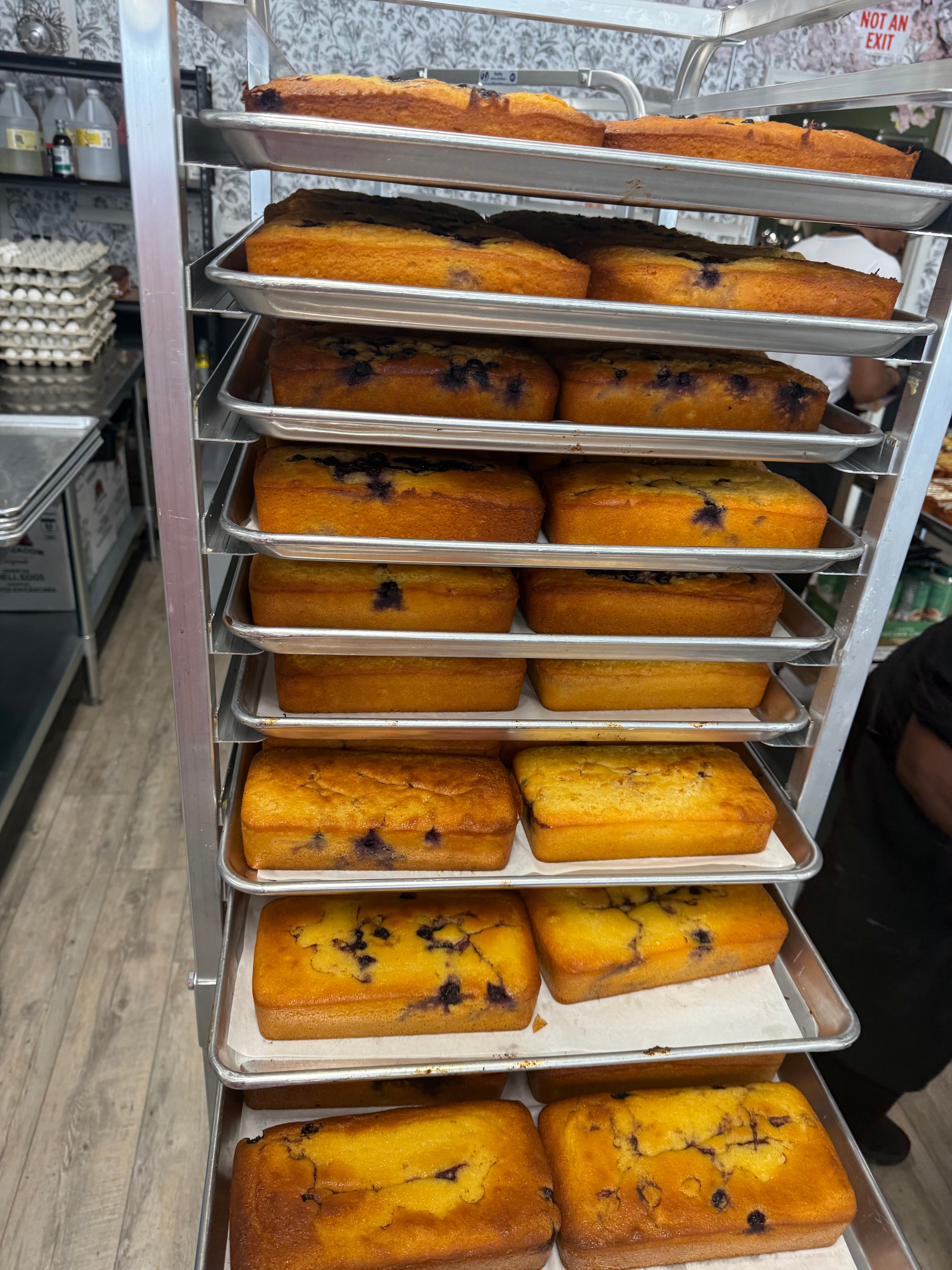 A metal bakery cart stacked with multiple trays of freshly baked blueberry bread loaves in a commercial kitchen setting.