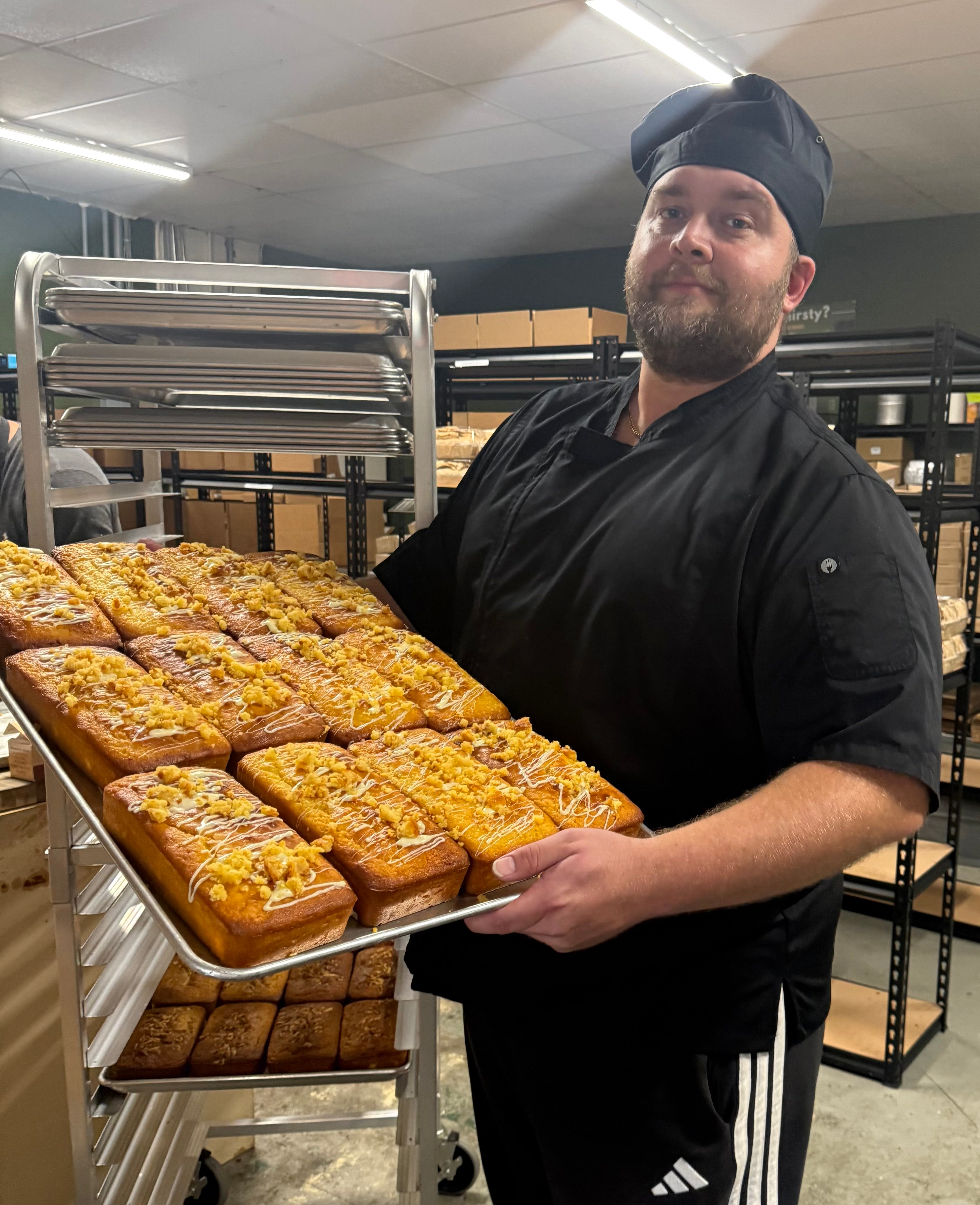 A baker in a black uniform and chef hat holds a large metal tray of freshly baked rectangular pastries topped with orange glaze in a bakery kitchen. Shelves with boxes and baking equipment are visible in the background.