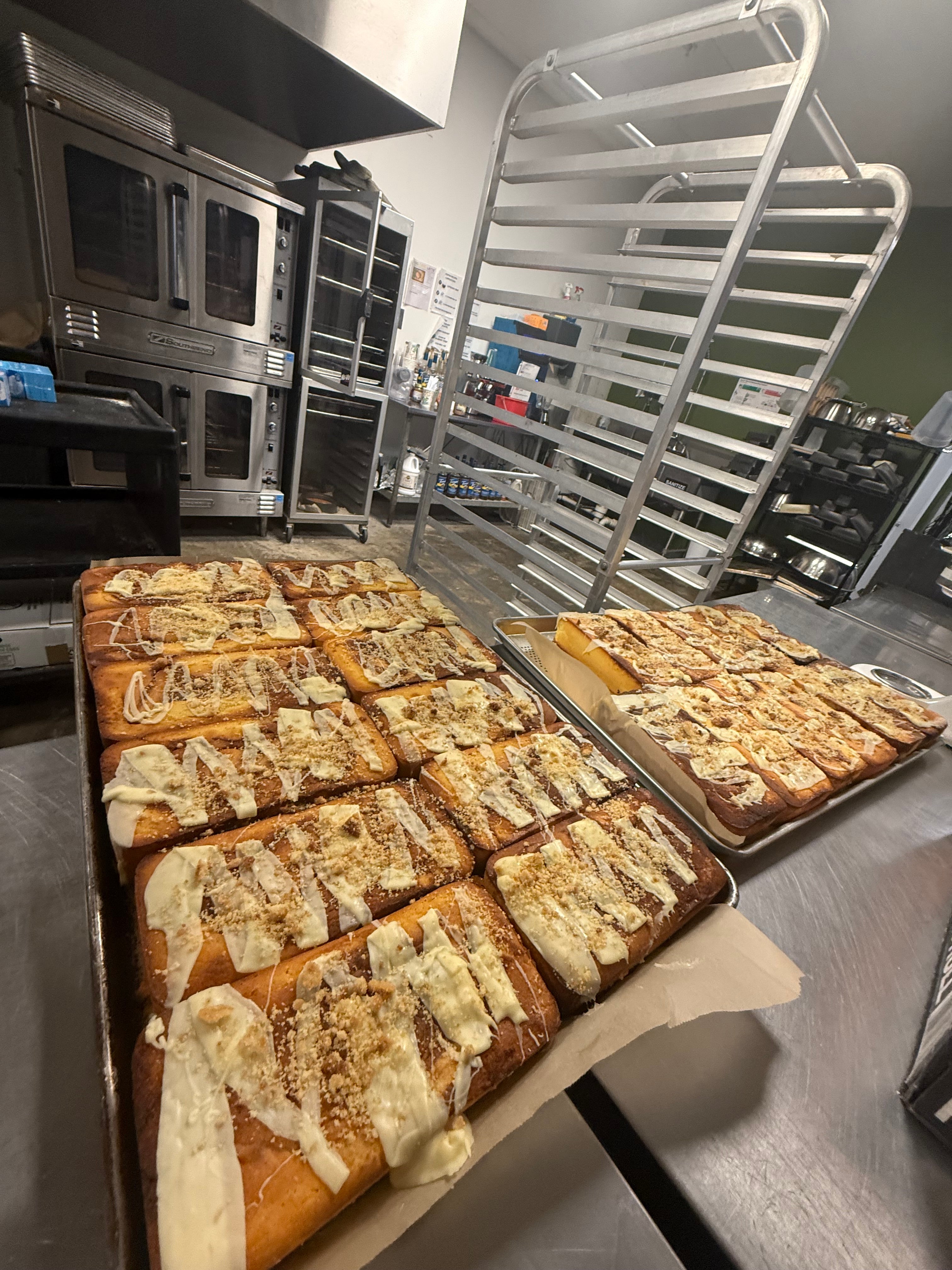 Two large trays of freshly baked cinnamon rolls topped with white icing and nuts, displayed in a commercial kitchen with stainless steel ovens and racks in the background.